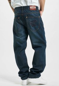Jeans de mezclilla azul oscuro con un corte relajado, que presentan una parche de cuero marrón en la cintura y un característico bordado del logo en el bolsillo trasero.