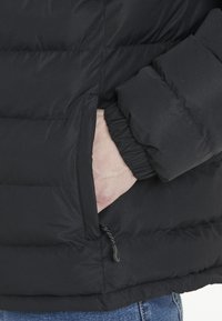 Chaqueta acolchada negra con textura de colcha, que cuenta con un bolsillo lateral y puños elásticos. Material suave con un brillo sutil.
