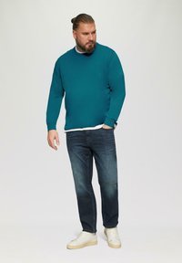 Tarnfarbener Pullover, Rundhalsausschnitt, lange Ärmel, kombiniert mit dunkelblauen Jeans und weißen Turnschuhen. Glatte Textur, lässiger Schnitt, minimale Nähtedetails.