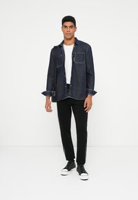 Jeans košulja s dugim rukavima u tamnoplavoj boji, kombinirana s bijelom majicom i crnim hlačama s bijelim bočnim prugama. Crne tenisice zaokružuju outfit.