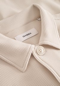 Kreemivärvi kraega särk, mis on valmistatud tekstureeritud kangast; omab nööpide sulgemist ja sildilt loetakse "GABBA."