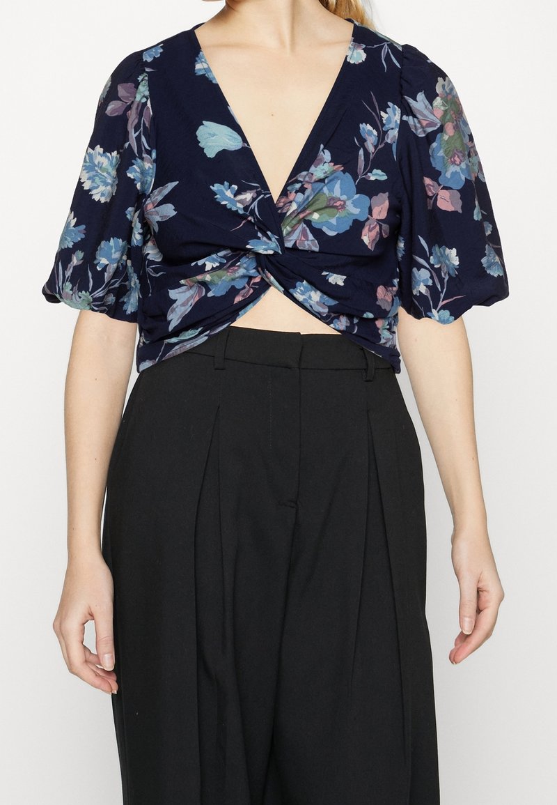 Femme portant un crop top bleu marine à fleurs à manches courtes avec un devant torsadé et un pantalon plissé noir taille haute.
