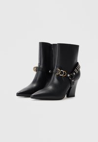 Bottines en cuir noir à bout pointu, avec une sangle décorative ornée d'anneaux dorés et argentés, et un talon bloc modéré.