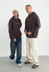 To personer har på seg store brune hettegensere med lommer foran og tekst, kombinert med casual jeans og kremfargede bukser. Bakgrunnen er nøytral.