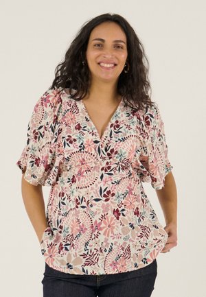 Lächelnde Frau mit dunklem lockigem Haar trägt eine hellrosa Bluse mit Blumenmuster und flatternden Ärmeln sowie dunkle Jeans, steht vor einem schlichten Hintergrund.