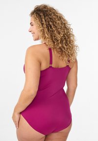 Magenta üheosaline ujumistrikoo õhukeste õlapaeltega, sileda tekstuuriga ja külgsete aktsendiga detailidega, mis näitavad keha järgivat selja- ja puusakujundust.