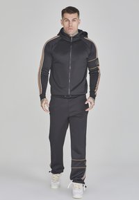 Zwarte zip-up hoodie met oranje strepen, gecombineerd met zwarte joggingbroeken met oranje accenten. Het materiaal lijkt soepel en rekbaar.