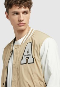Jachetă bomber bej cu un tricou alb cu mânecă lungă. Prezintă un "A" brodat și "GENUINE" pe piept, cu un guler contrastant cu dungi.