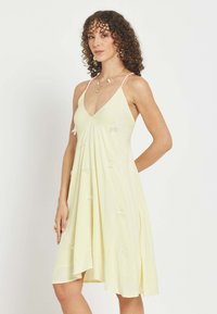 Mujer con cabello rizado que lleva un vestido sin mangas de color amarillo pálido con delicados detalles florales blancos, de pie frente a un fondo blanco liso.