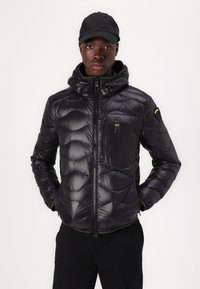 Giubbotto imbottito nero con design trapuntato, chiusura a zip e cappuccio. Presenta una tasca sul petto e accenti verdi sull'orlo. Finitura opaca e materiale leggero.