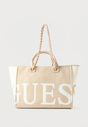 Sac cabas en toile beige et blanche avec logo « GUESS » en grandes lettres blanches et anses tressées avec détails en chaîne dorée.