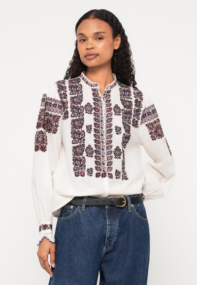 Fehér, hosszúujjú ing, bonyolult bordó és színes virágmintákkal hímezve. Gombos gallér, részletes mandzsettákkal. Kék farmerral viselve.