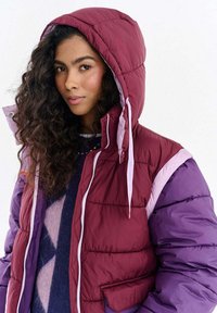 Kurtka puchowa w kolorach bordowym, fioletowym i różowym w formie bloków kolorystycznych. Posiada kaptur, zamek błyskawiczny i pikowaną fakturę.