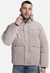 Beige corduroy pufferjack met een hoge kraag, drukknoopsluiting en twee voorzakken. De stof heeft een verticale ribbelstructuur.