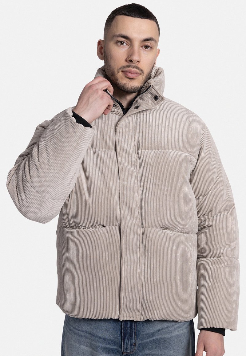 Beige corduroy pufferjack met een hoge kraag, drukknoopsluiting en twee voorzakken. De stof heeft een verticale ribbelstructuur.