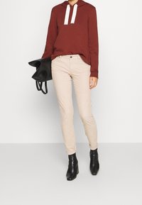 Maroon hooded sweatshirt with white drawstrings, paired with beige skinny jeans and black heeled ankle boots. Black bag held in one hand.