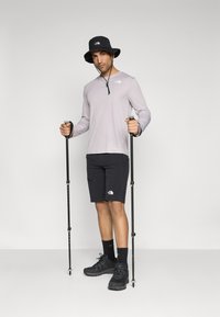 Hele lilla pikad seljae, mustad lühikesed püksid logoga, must müts, mustad matkajalatsid, hoides matkakeppe, seismel lihtsal taustal.