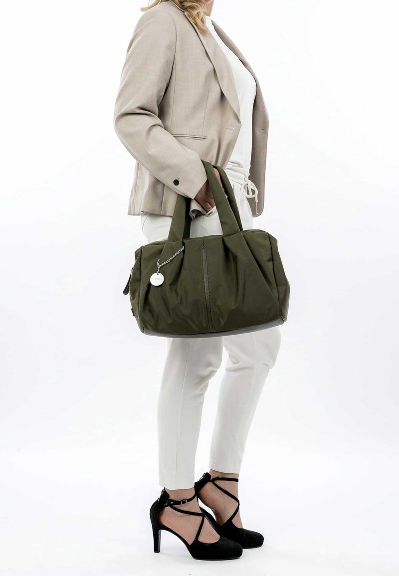 Femme tenant un grand sac à main vert olive, portant un blazer beige, un pantalon blanc et des chaussures à talons noirs avec des brides à la cheville.