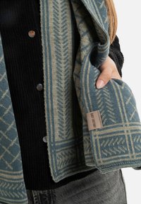 Geweven sjaal in blauw en beige met geometrische patronen en een gestructureerde afwerking. Bevat een label aan de zijkant en metalen knopen voor bevestiging.