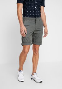 Uomo che indossa pantaloni corti grigio scuro arrotolati, una camicia blu navy a maniche corte con quadrati bianchi e sneaker bianche su sfondo neutro.