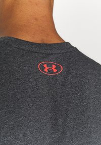 Dos de la nuque d'une personne et chemise en tissu gris avec un petit logo rouge Under Armour près de l'encolure.