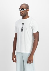 Hvid bomulds t-shirt med et vertikalt sort "ALPHA" grafik på forsiden. Modellen bærer reflekterende solbriller og lys blå jeans.