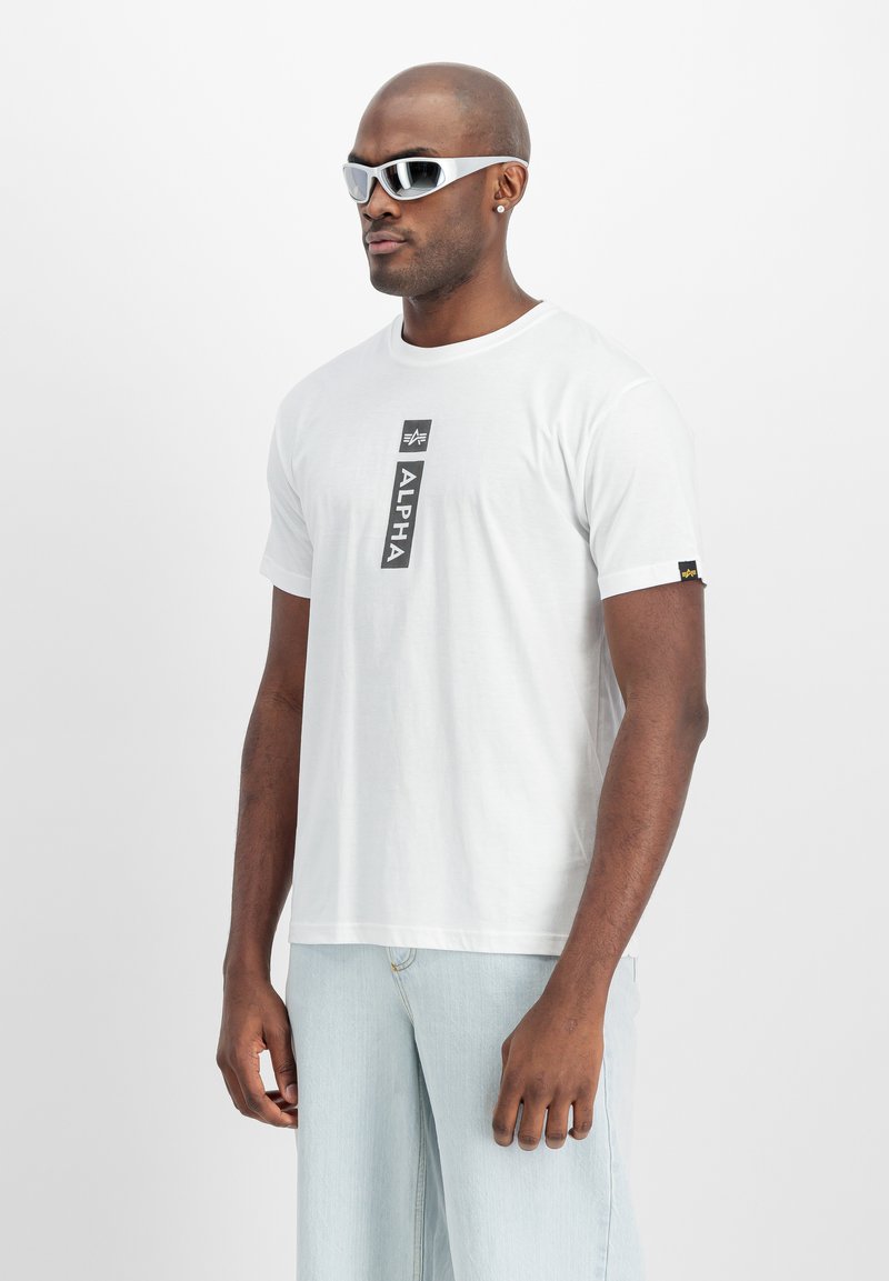 Hvid bomulds t-shirt med et vertikalt sort "ALPHA" grafik på forsiden. Modellen bærer reflekterende solbriller og lys blå jeans.