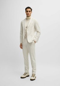 Costume beige clair avec un blazer ajusté et un pantalon slim, associé à un col roulé crème et des bottes à lacets beiges. Détails en tissu texturé visibles.