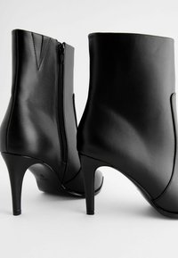 Melna ādas potītes zābaki ar elegantu apdari, augstiem stiletto papēžiem un sānu rāvējslēdzējiem. Satur smalkas šuvju detaļas uz apakšdaļas.