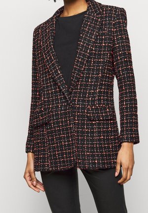 Vrouw draagt een zwarte tweedblazer met een roze-wit geruit patroon, gecombineerd met een zwart shirt en zwarte broek.