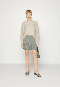 Frau in einem beigefarbenen, flauschigen Pullover, grauem, gefälteltem Minirock, grauen, hohen Sandalen, hält eine kleine graue Handtasche und steht vor einem einfarbigen Hintergrund.