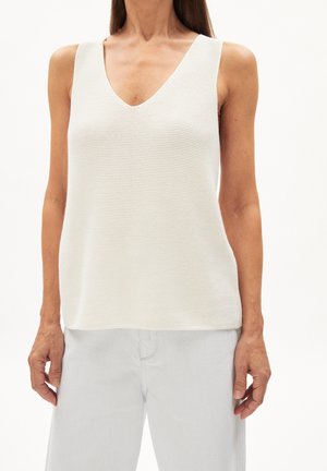 Femme portant un haut en maille blanc sans manches avec un col en V et un pantalon de couleur claire, vue du cou aux hanches sur un fond uni.