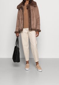 Brun mockajacka med pälskrage, dragkedja och metallaccenter. Beigetröja, krämfärgade smalbyxor och ljusrosa slip-on sneakers. Svart handväska.