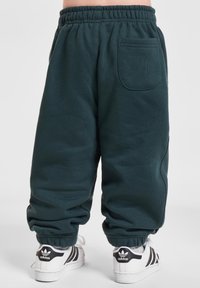 Dunkelblaugrüne Jogginghose mit elastischem Bund, lockerer Passform, Bündchen an den Knöcheln und einer Gesäßtasche, kombiniert mit weißen Sneakers mit schwarzen Streifen.