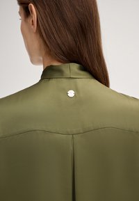 Blusa di seta verde oliva con colletto alto e dettaglio di bottone tondo argentato sul retro, con una texture liscia e linee pulite.