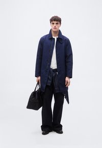 Νεαρός άνδρας φοράει μπλε σκούρο παλτό πάνω από λευκό πουκάμισο με σκούρο παντελόνι και μαύρα παπούτσια, κρατώντας μαύρη τσάντα, στέκεται μπροστά σε μια απλή ανοιχτή φόντο.