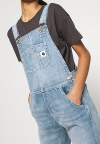 Lichtblauwe denim overall met verstelbare banden, metalen hardware, een voorkleedzak en zijzakken, gedragen over een donker t-shirt.