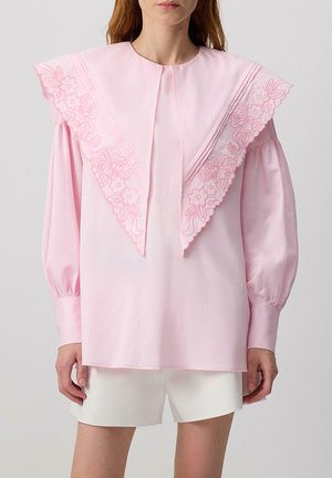 Blusa rosa con un ampio colletto ricamato con motivi floreali, maniche lunghe e polsini arricciati, abbinata a pantaloni corti bianchi.