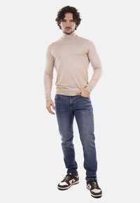 Maglione beige a collo alto con design aderente, abbinato a jeans blu e sneaker marroni e bianchi, indossato da un modello maschile.