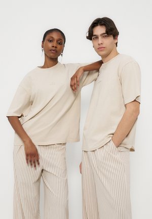 Dvaja modeli oblečení v béžových nadrozmerných tričkách a béžových širokých nohaviciach s prúžkami, stojiaci pred jednoduchým bielym pozadím.