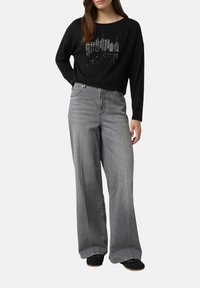 Frau trägt ein schwarzes Langarmshirt mit Druck der New Yorker Skyline und hellgraue weite Jeans und steht vor einem weißen Hintergrund.