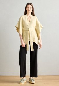 Vêtement beige en style cache-coeur avec des manches courtes et une ceinture à nouer. Porté sur un pantalon noir et des chaussures grises. Texture lisse, design minimal.
