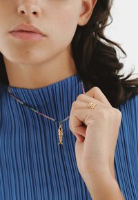 Colgante de pez dorado en un cordón multicolor, llevado con una blusa azul con pliegues. La mano muestra un anillo de textura dorada.