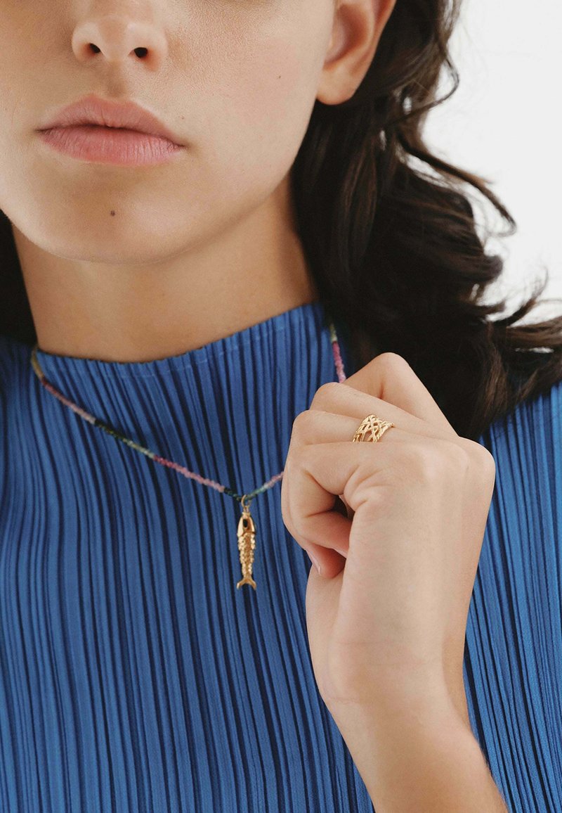 Colgante de pez dorado en un cordón multicolor, llevado con una blusa azul con pliegues. La mano muestra un anillo de textura dorada.