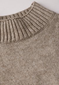 Pull en tricot beige avec un col côtelé, présentant une texture douce et des variations subtiles de couleur à travers le tissu.
