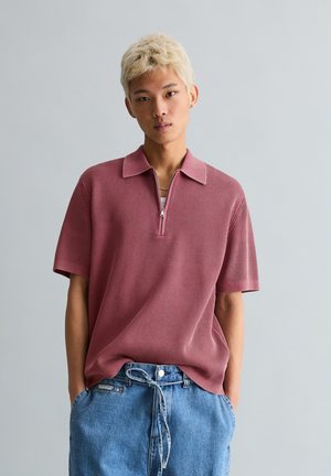 Junge Person mit kurz gebleichtem Haar, die ein staubrosa geripptes Poloshirt und lockere blaue Jeans trägt, mit Händen in den Taschen.