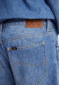 Jeans in denim azzurro chiaro con un'etichetta in pelle marrone con la scritta "Lee". Dettagli di cucitura e design delle tasche ben visibili.