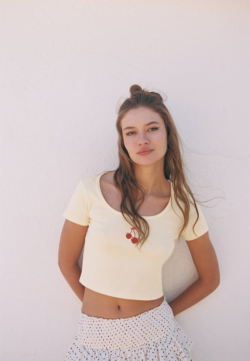 Gul crop top med rund halsringning och en röd körsbärsdesign. Kombinerad med en vit kjol prydd med svarta prickar.