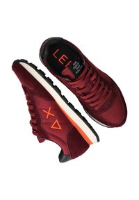 Un paio di sneakers bordeaux con loghi geometrici arancioni sui lati, suole nere con tasselli e il testo "CORRI PIÙ VELOCE DI QUANTO PUOI" sull'etichetta interna della linguetta.