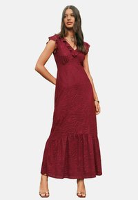 O femeie îmbrăcată într-o rochie maxi burgundy fără mâneci, cu decolteu în V și margine volanată, stând cu o mână pe șold, purtând tocuri și un colier.
